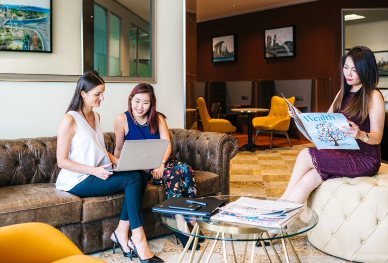 Three women in a stylish lounge working on a laptop and reading, surrounded by modern decor and cozy seating