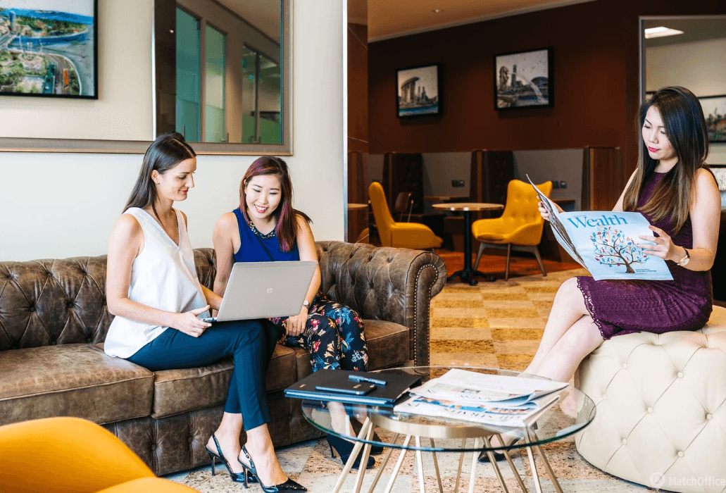 Three women in a stylish lounge working on a laptop and reading, surrounded by modern decor and cozy seating