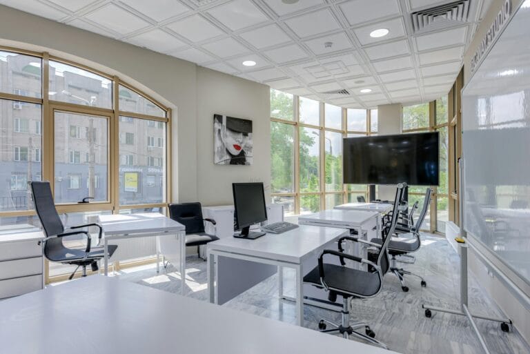 Modern office with bright windows, desks, and a large screen for presentations.