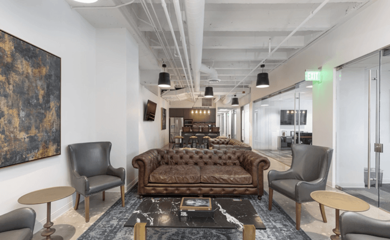 Cozy office lounge with brown leather sofa, gray armchairs, modern decor, and glass-walled offices nearby
