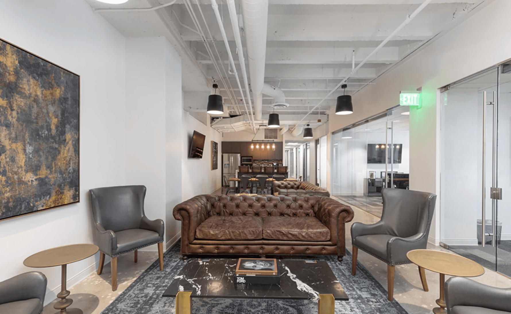 Cozy office lounge with brown leather sofa, gray armchairs, modern decor, and glass-walled offices nearby