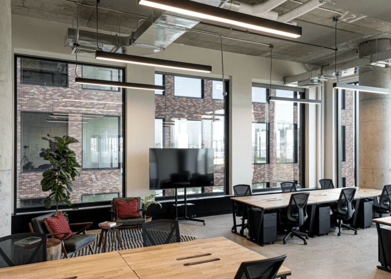 Modern coworking office with wood desks, ergonomic chairs, large windows, and industrial ceiling lights