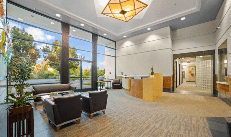 Bright office lobby with modern furniture and large windows showing outdoor greenery.